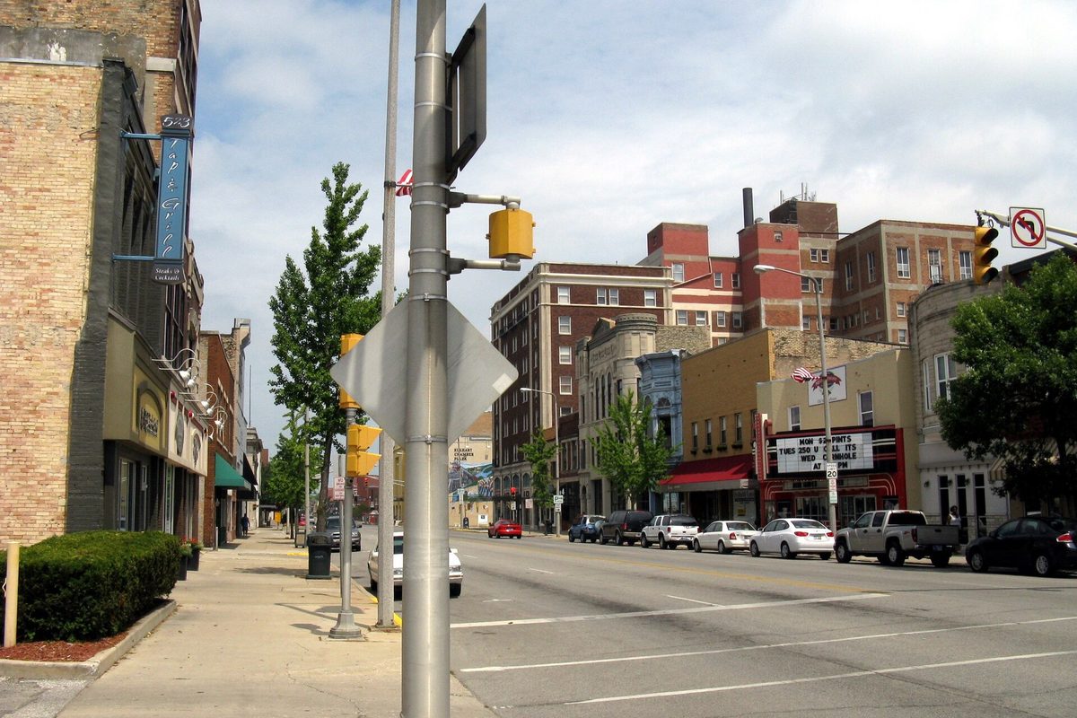 Main Street in downtown Elkhart looking north, with brick storefronts, a theater marquee, traffic signals and parked cars and pickups