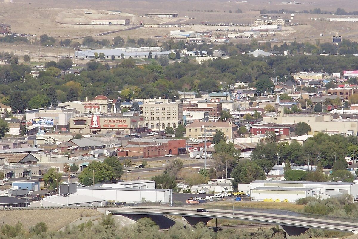 Downtown Elko seen from a bluff to the south, with low brick buildings, a red casino sign, green trees, a highway bridge in front and dry hills behind