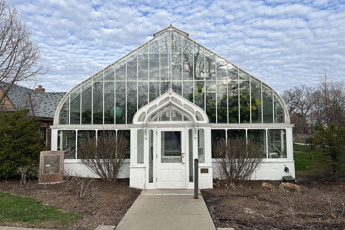 The white-framed glass Wilder Park Conservatory in Elmhurst, with a peaked roof, arched entry and bare shrubs under a sky of patchy clouds