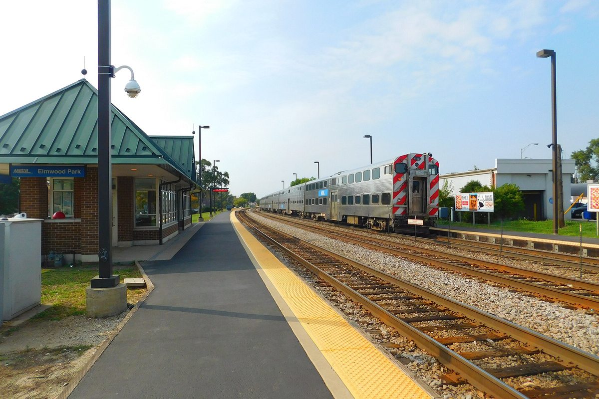 The Elmwood Park Metra station, a small brick shelter with a green metal roof beside the platform, with a silver Metra train on the tracks