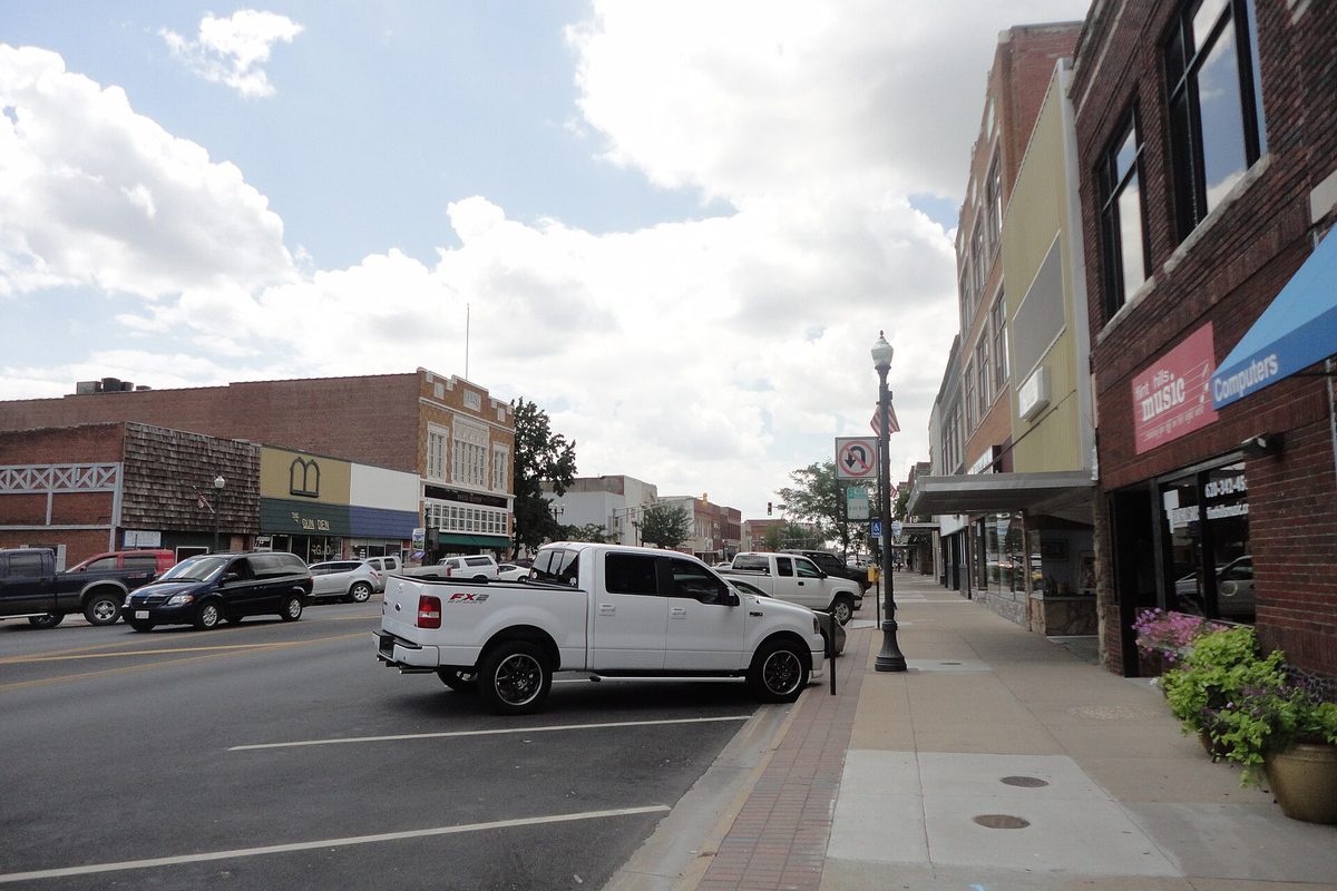 A white pickup parked at the curb on a downtown Emporia street lined with brick storefronts, shop signs and flower planters under a cloudy sky