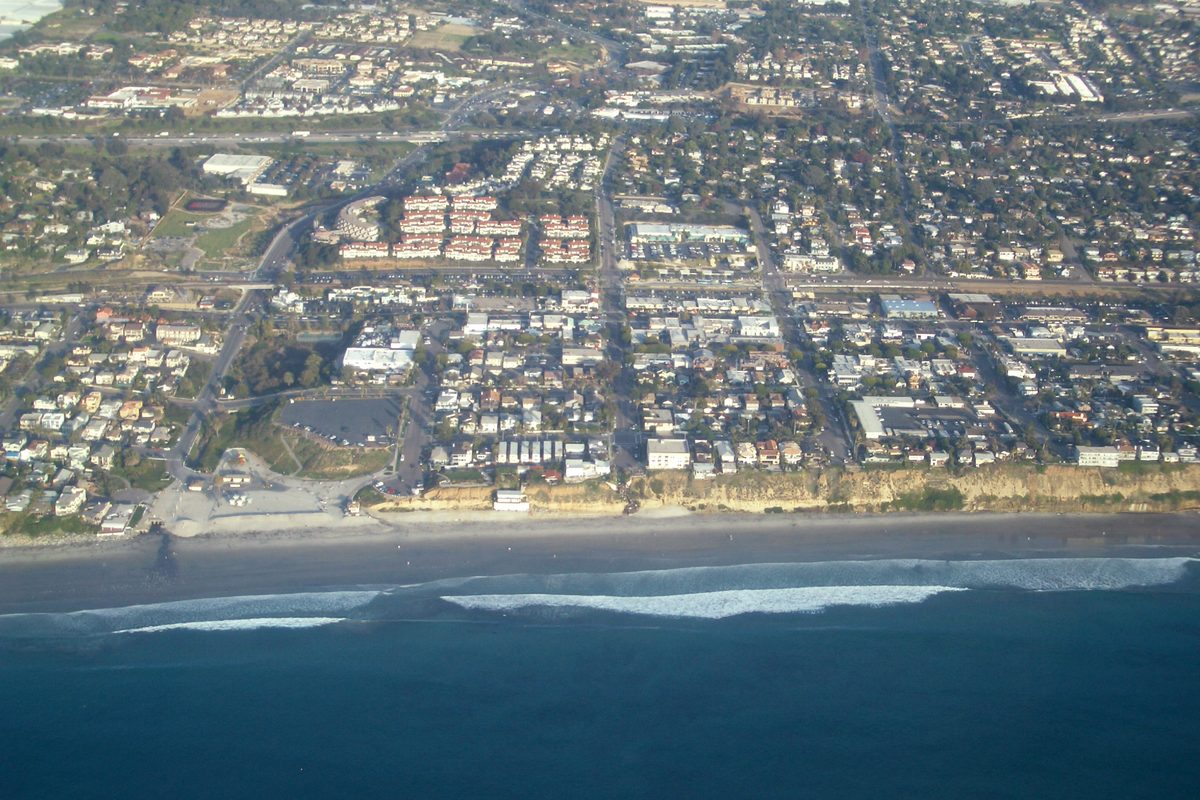 Aerial view of Encinitas from offshore: waves rolling onto the beach below low bluffs, with neighborhoods spreading inland