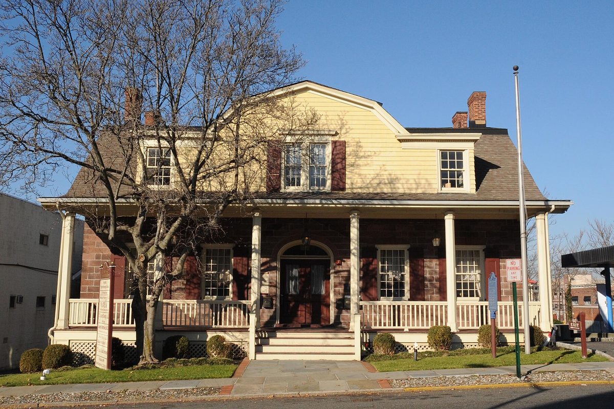 The John G. Benson House in Englewood, a yellow Dutch Colonial with a gambrel roof, red shutters, brick lower walls and a white columned porch, under a clear blue sky