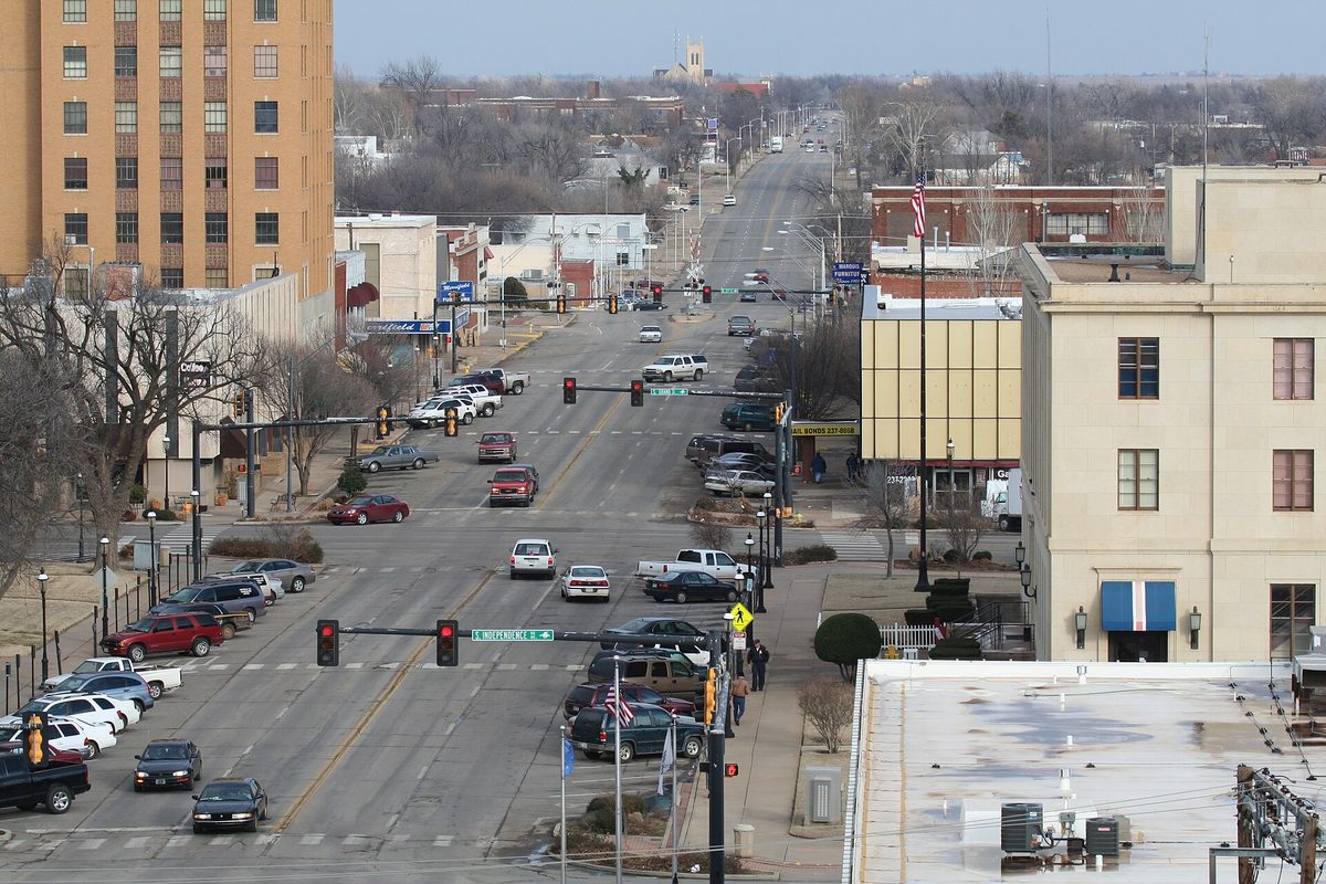 Looking east down Broadway Avenue in Enid from above, with traffic lights, cars parked along the curbs, a tall tan building on the left and bare winter trees
