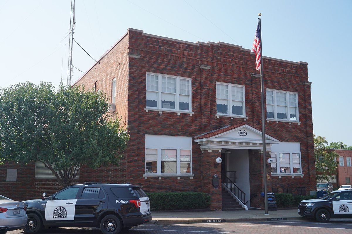 Ennis City Hall, a two-story red-brick building with a date stone over the door and a flagpole, with police SUVs parked along the curb