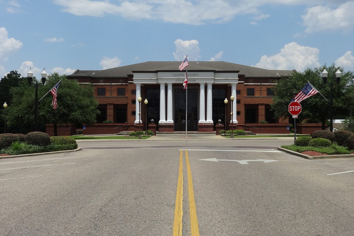 The Coffee County Courthouse in Enterprise, a brick building with a white-columned portico and American flags, at the end of a drive with a stop sign