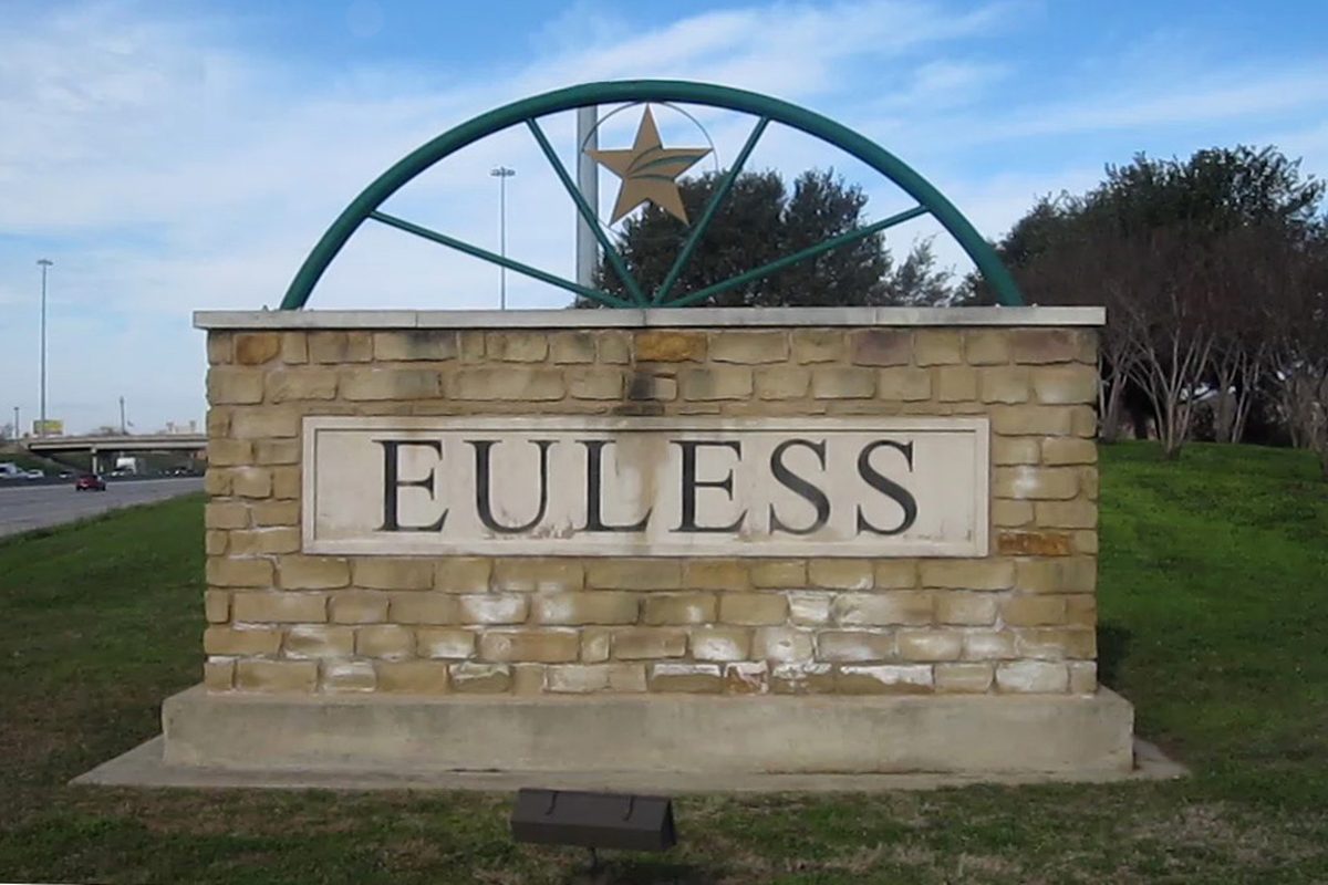 Stone Euless welcome sign under a green metal arch with a Texas star, standing on a grassy slope beside a highway