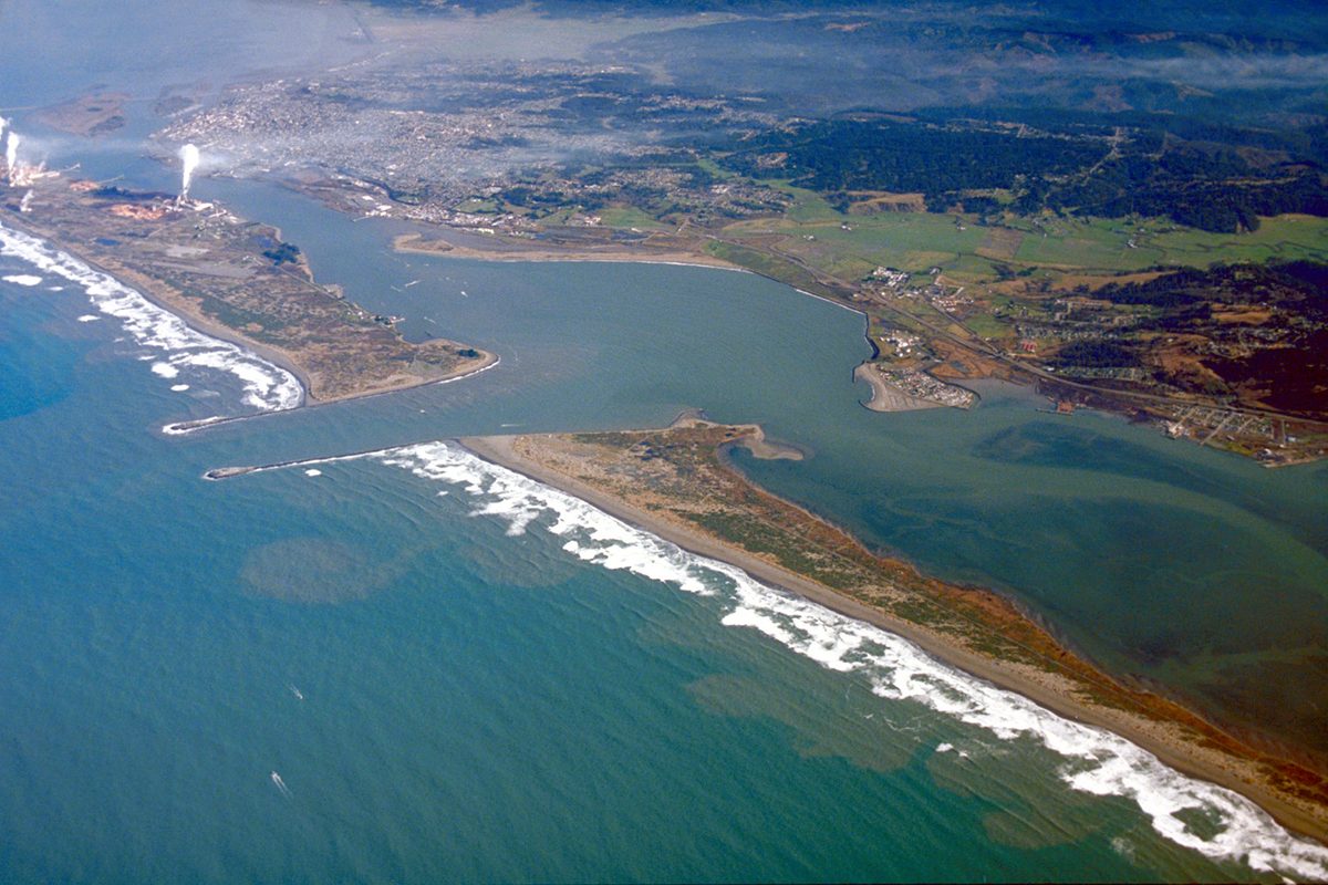 Aerial view looking northeast over the mouth of Humboldt Bay, with ocean surf on two sand spits and the city of Eureka spread along the far shore