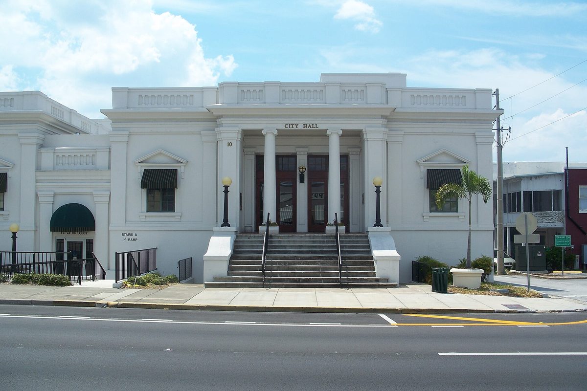 Eustis City Hall, a white neoclassical building with columns, a central stairway, lamp posts and a palm tree, seen across the street