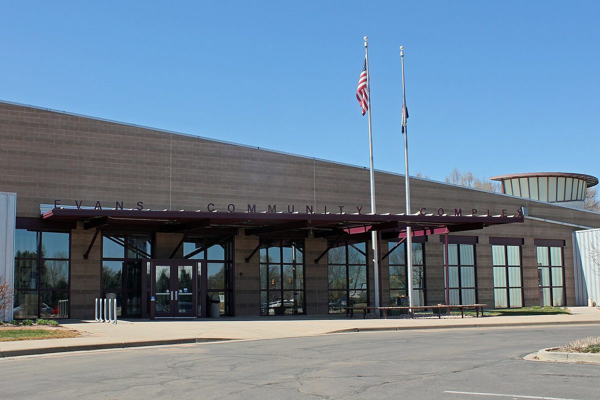 The long, low Evans Community Complex, with Evans Community Complex lettered over a glass entrance and two flagpoles out front under a clear sky