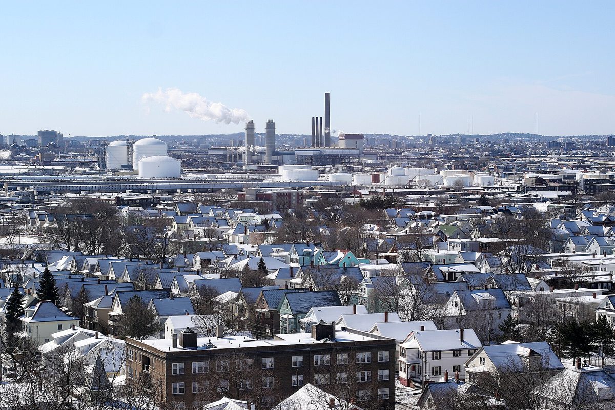 Snow-covered rooftops of closely packed houses in Everett, with white storage tanks and smokestacks behind them under a pale winter sky