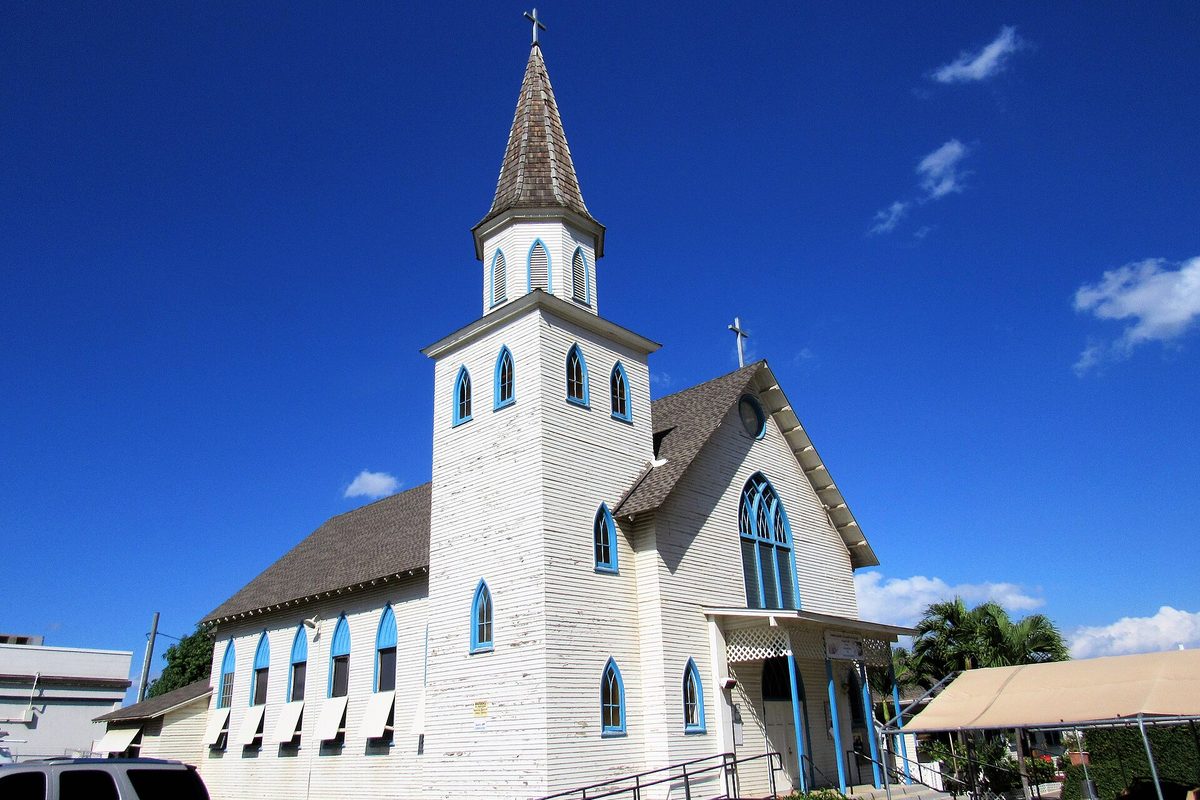 Immaculate Conception Catholic Church in Ewa, a white wooden church with blue-trimmed arched windows and a tall shingled steeple, under a deep blue sky