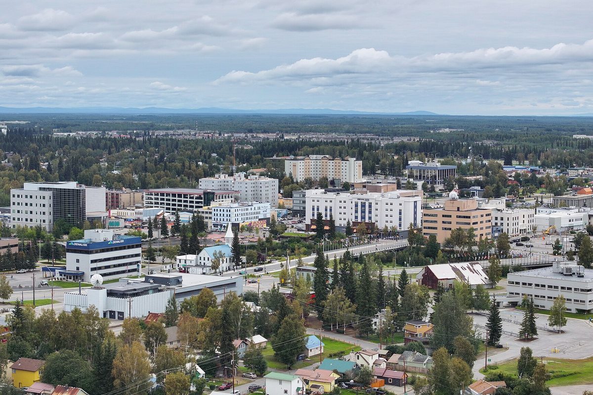 Aerial view of downtown Fairbanks in summer, with mid-rise buildings, a bridge and small houses among green trees under a cloudy sky