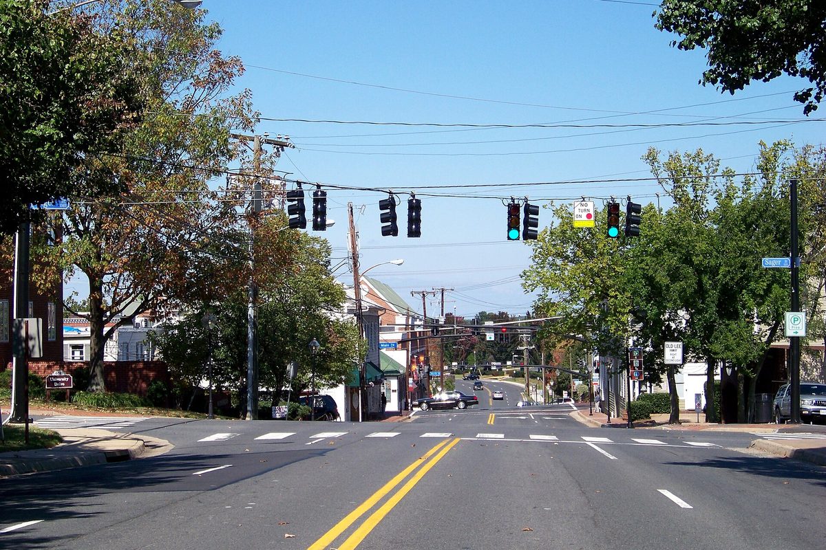 A four-lane street in Fairfax with double yellow center lines and traffic lights strung overhead, trees and low brick buildings on both sides under a clear blue sky