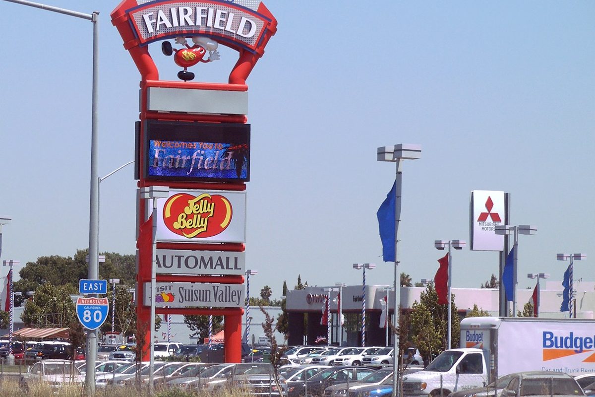 A tall red Fairfield welcome sign beside Interstate 80, with a digital board and an auto mall lot full of parked cars behind it