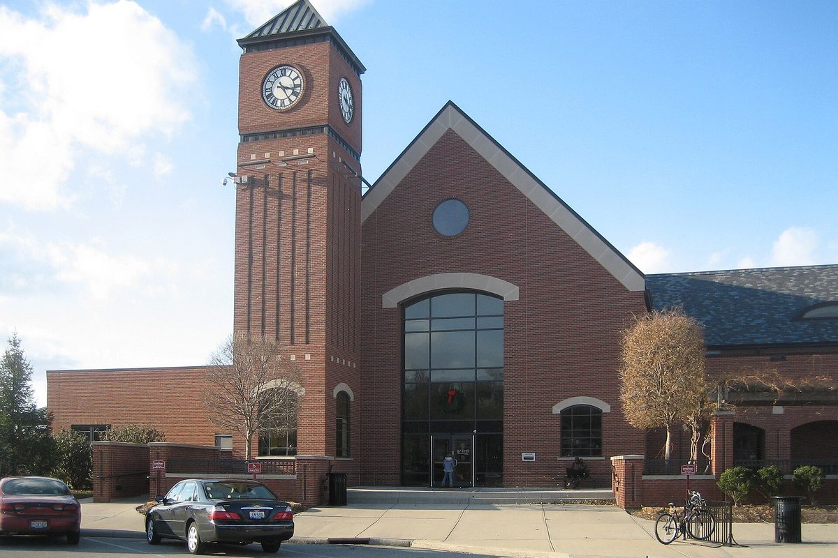 The Fairfield public library, a red brick building with a tall clock tower and an arched glass entrance, with cars parked out front on a sunny day