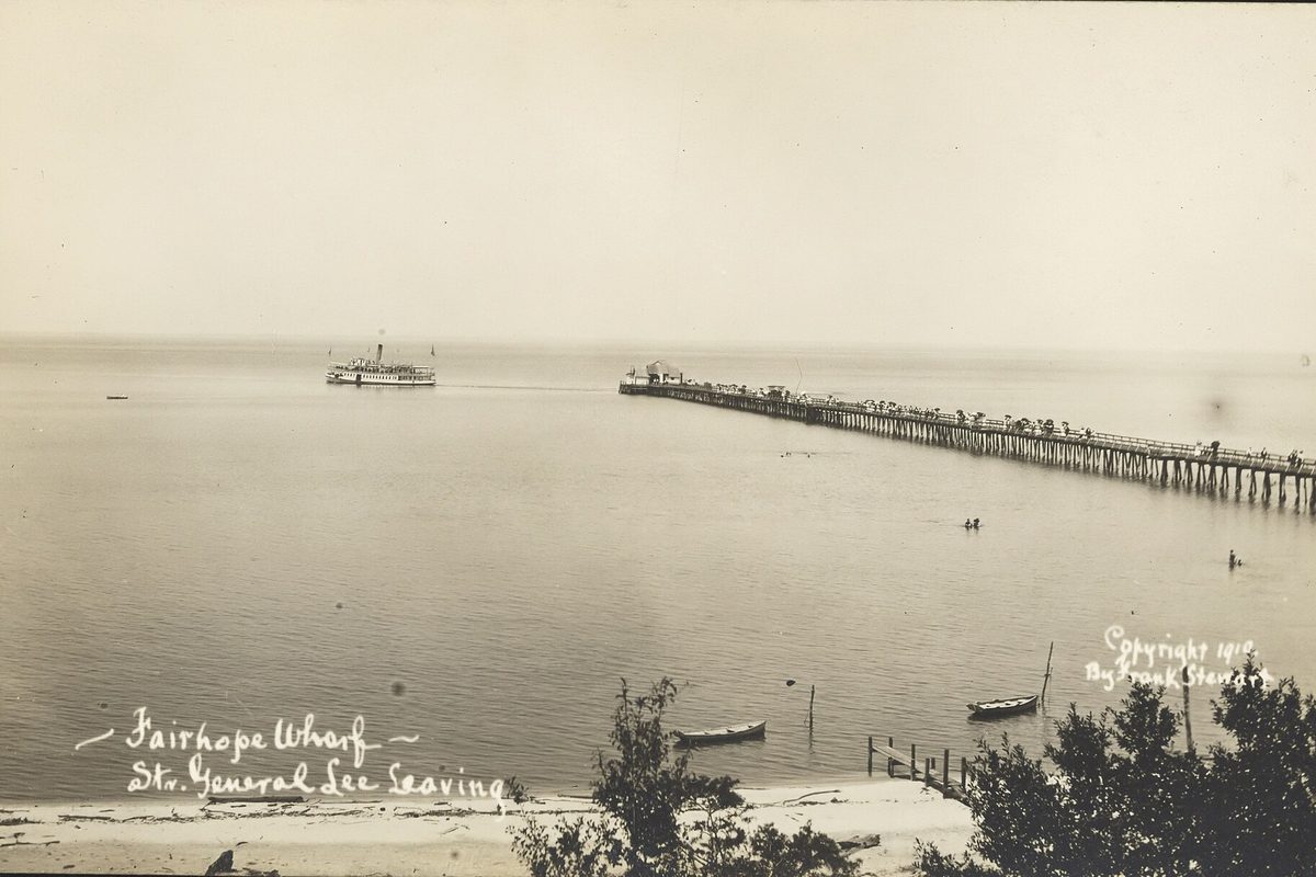 A 1910 sepia photograph of the long Fairhope Wharf reaching into Mobile Bay as the steamer General Lee pulls away, small boats on the beach below