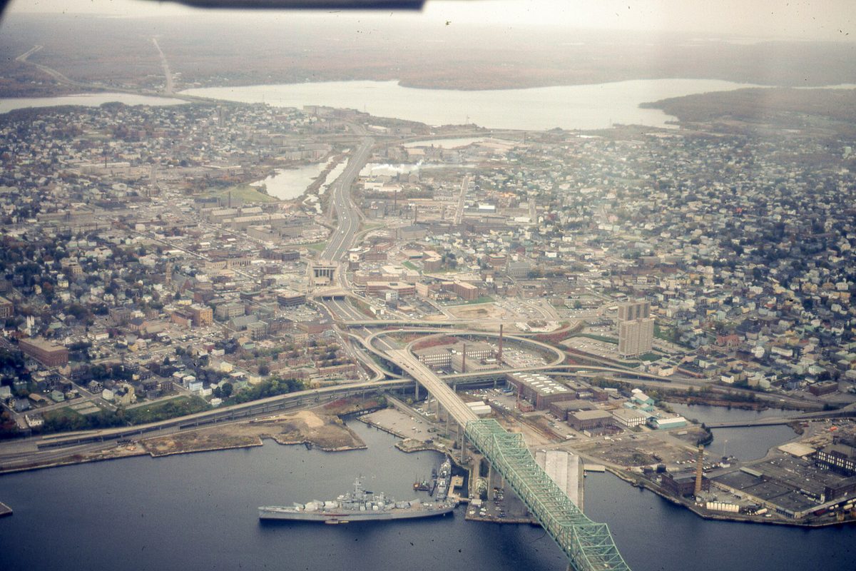 Aerial view of Fall River's packed neighborhoods and highway ramps, with a long green truss bridge over the water and a gray naval ship docked below