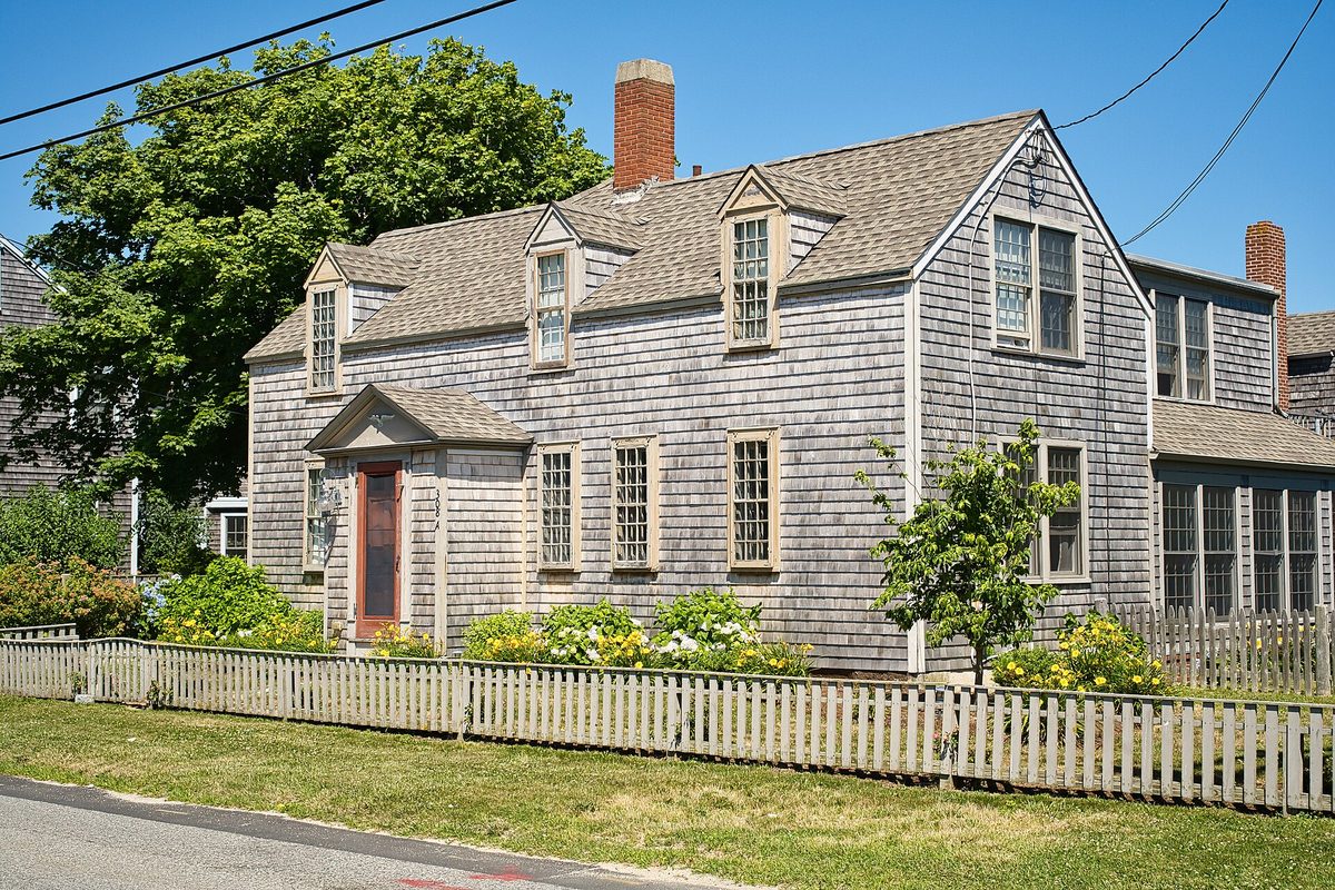 A gray-shingled Cape house with a red front door and a weathered picket fence at 308 Shore Street in Falmouth, under a clear blue sky