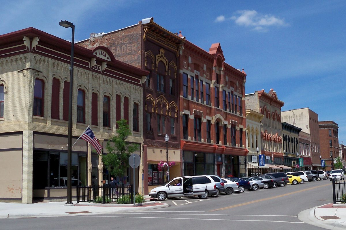 A row of ornate red and cream brick storefronts on a downtown Faribault corner, with an American flag and cars parked along the street