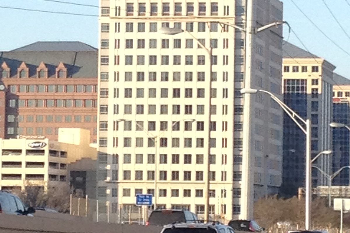 A tall pale office tower with rows of small windows beside other mid-rise buildings, seen from a highway lined with street lights