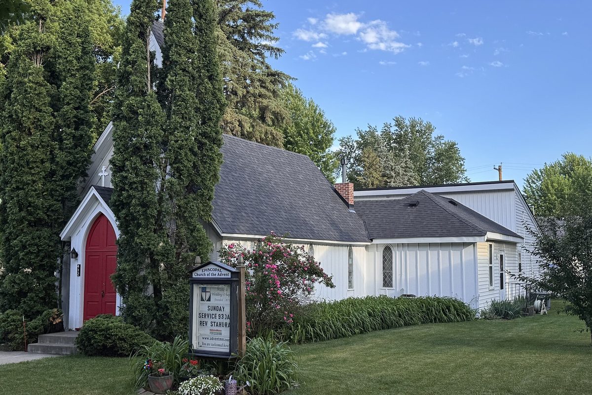 The white Episcopal Church of the Advent in Farmington, with a red arched door, tall evergreens and a pink flowering shrub out front