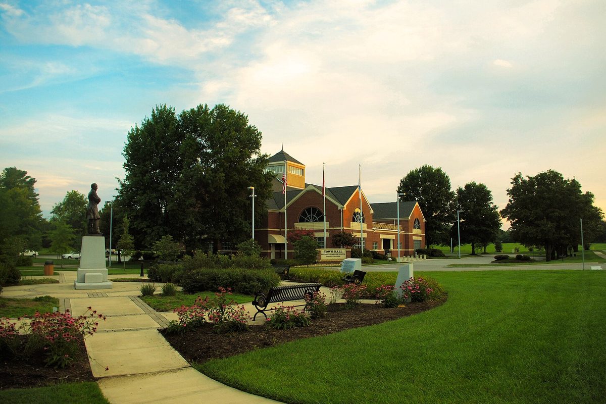 Farragut Town Hall, a brick building with a cupola and flags, behind a green lawn, flower beds, a bench and a statue on a stone pedestal