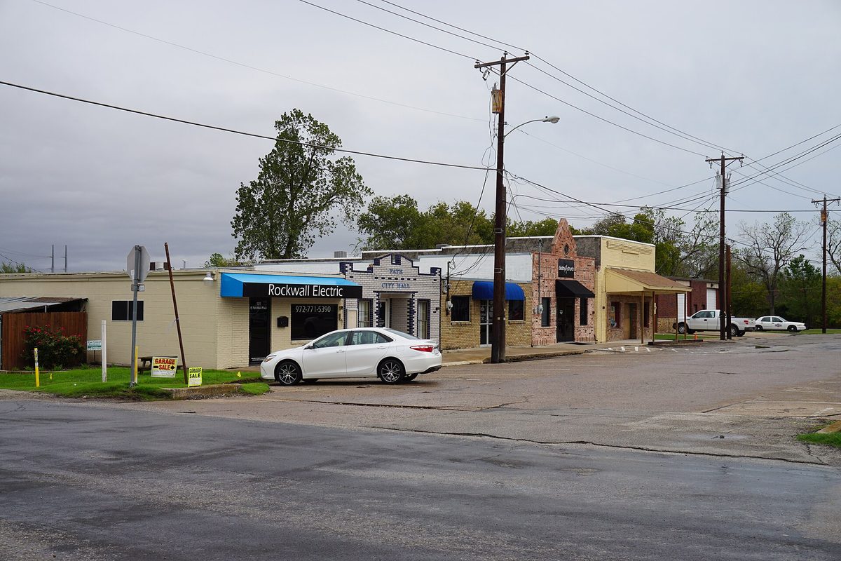 Old one-story storefronts on Fate Main Place, with a white sedan parked out front and power lines overhead on a cloudy day