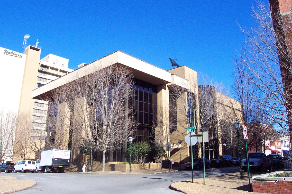 The tan University of Arkansas Center for Continuing Education building in Fayetteville, with bare trees in front and a hotel tower behind it under a clear blue sky