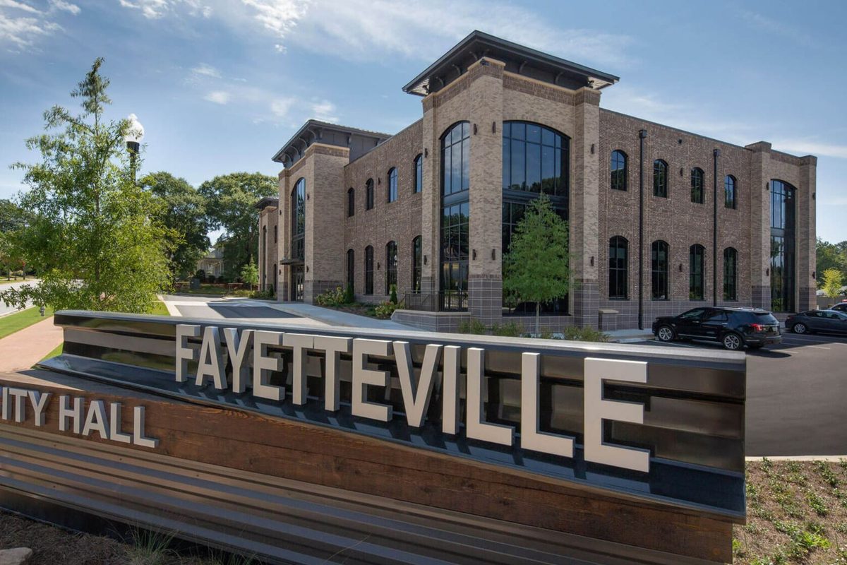 A Fayetteville City Hall sign with large silver letters in front of a two-story brick building with tall arched windows and a parking lot