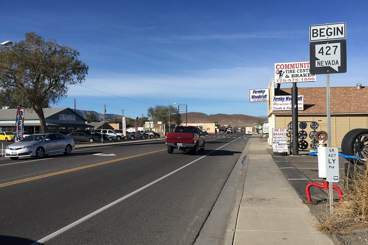 Main Street in Fernley looking west, with a Begin 427 Nevada route sign, a tire and brake shop, a red pickup on the road and brown hills under a clear blue sky
