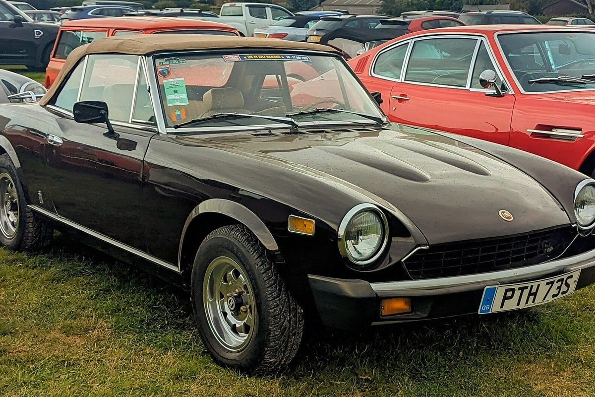 A dark brown original-style Fiat 124 Spider roadster with a tan soft top and round headlights, parked on grass at a car show