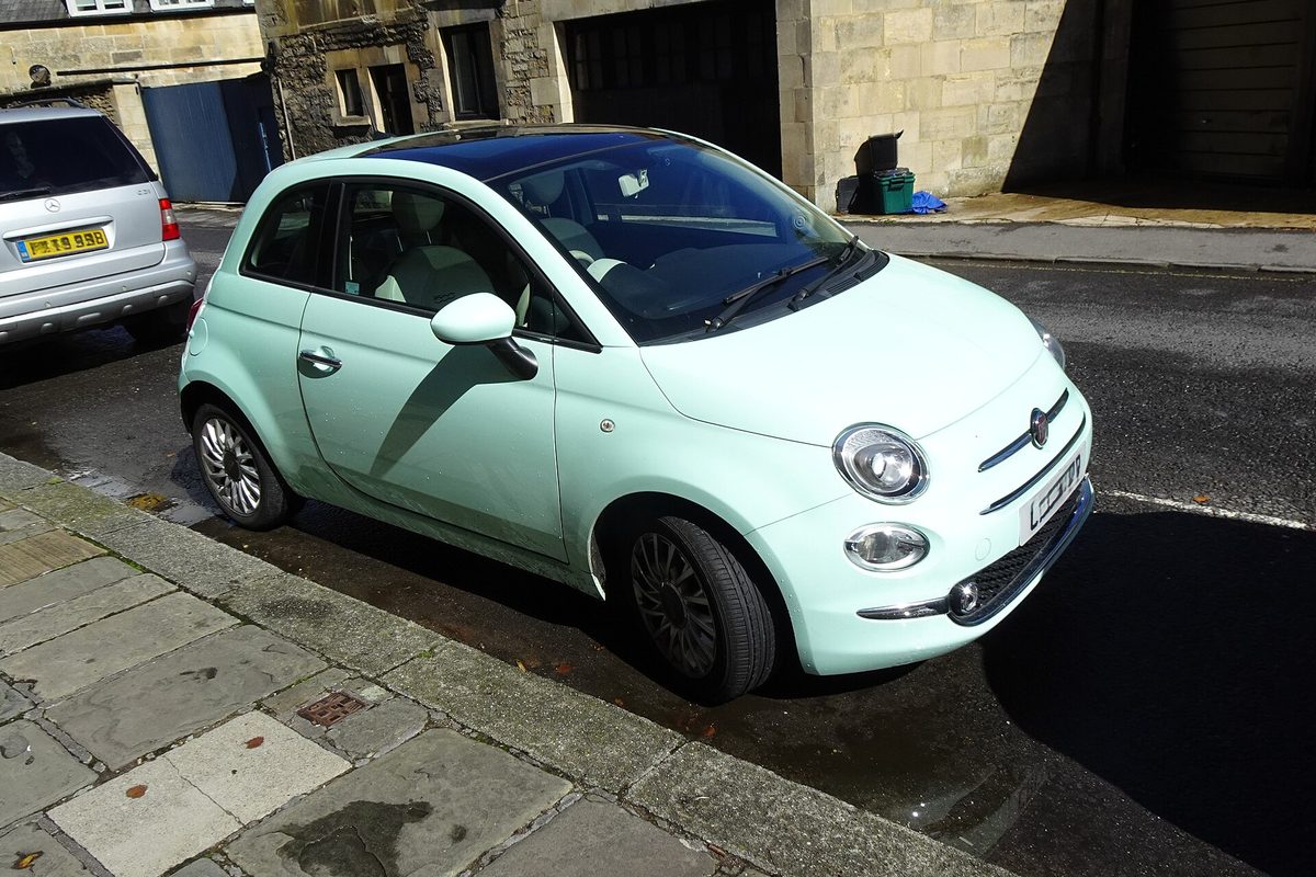 A mint-green Fiat 500 hatchback with a dark glass roof, parked at the curb on a stone-paved street