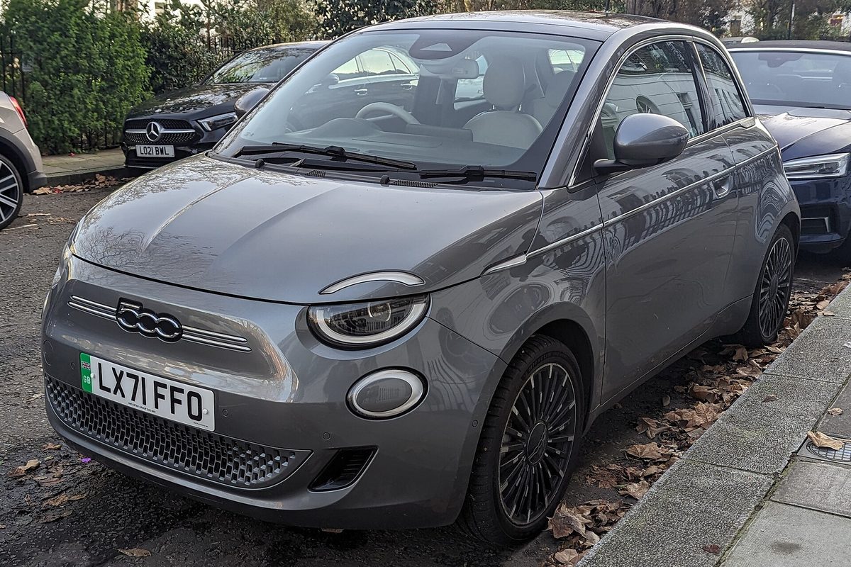A gray third-generation Fiat 500e hatchback with oval LED headlights, parked at a curb in fallen autumn leaves