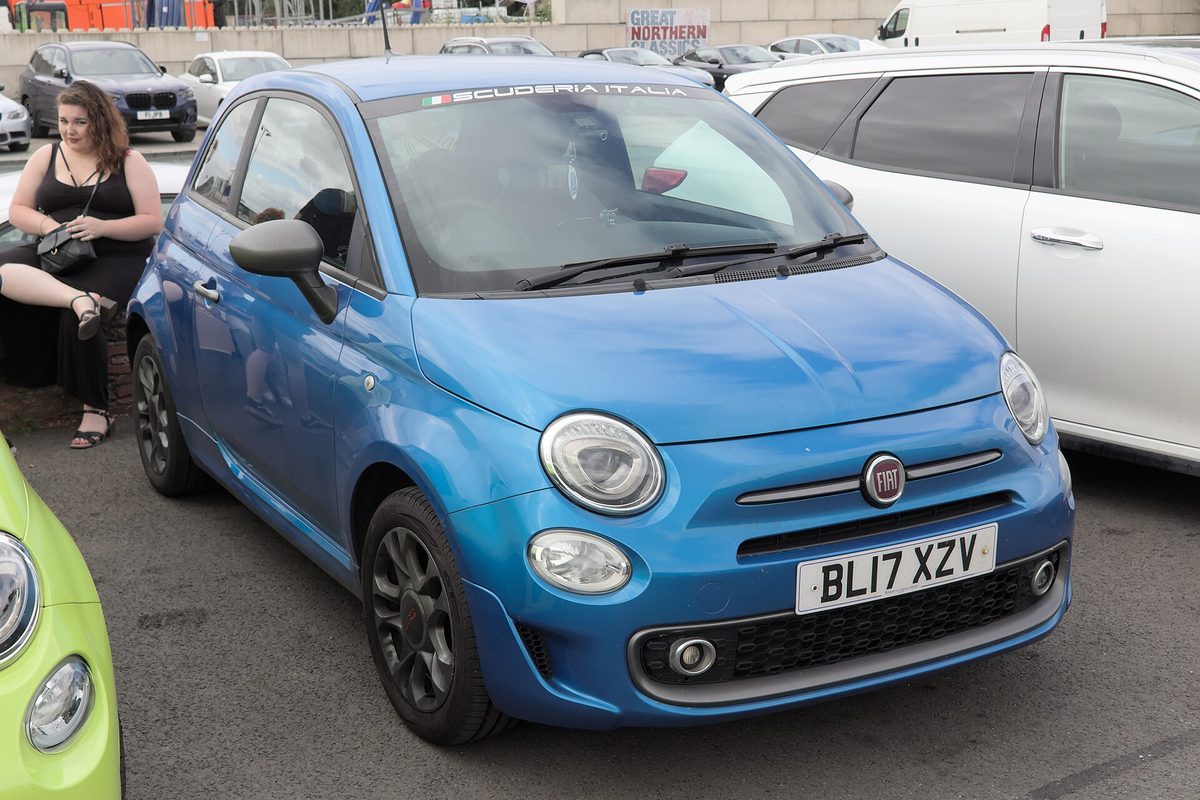 A bright blue Fiat 500 hatchback parked in a lot among other cars at the Great Northern Classics show, with a lime-green car at the left edge