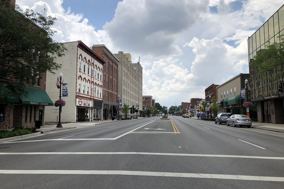 Main Street in downtown Findlay on a summer day, with brick and stone storefronts, flower baskets on the lamp posts and a wide, nearly empty road