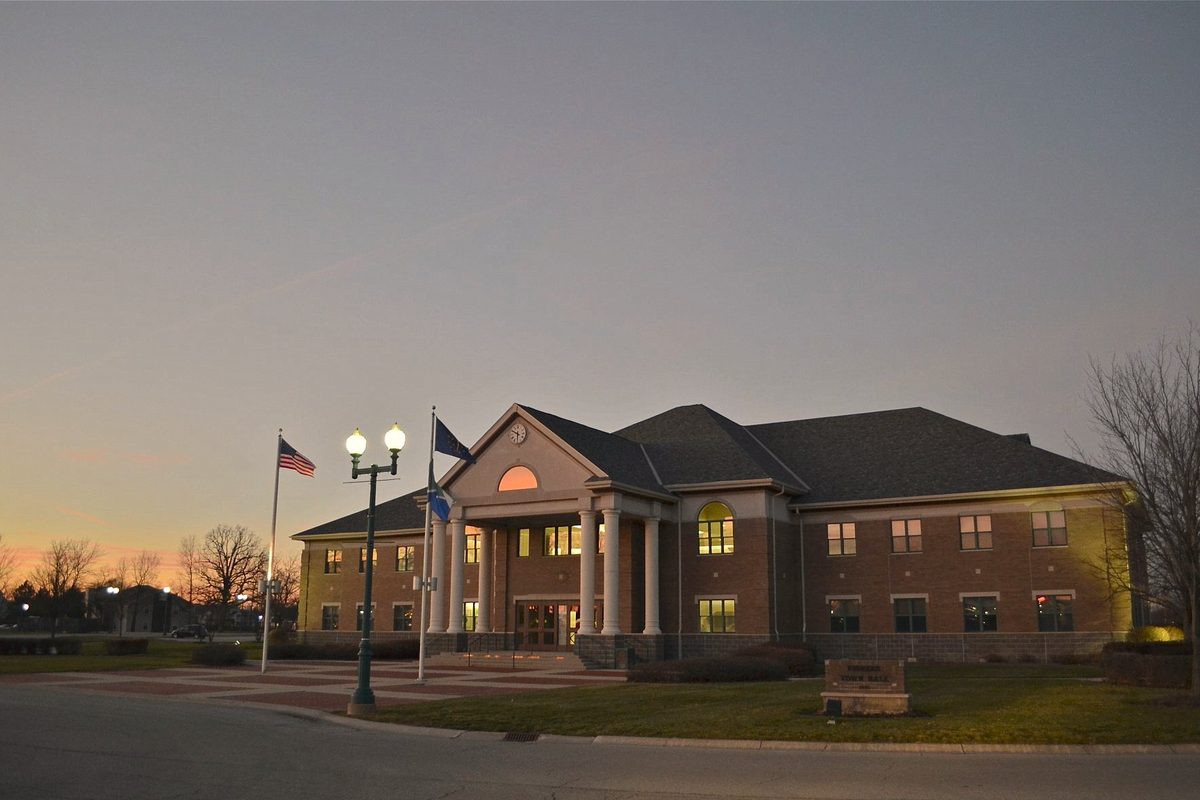 Fishers City Hall, a brick building with a white-columned entrance and a lit clock in the gable, at dusk with flags out front