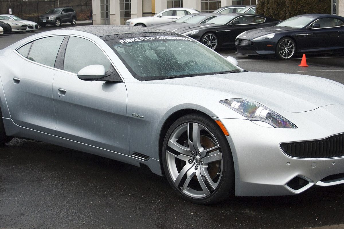 A silver Fisker Karma sedan on a wet dealer lot, with dark sports cars parked in a row behind it
