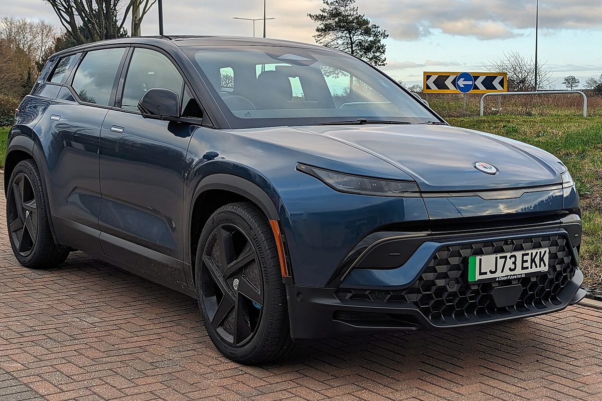 A dark blue Fisker Ocean electric SUV parked on a brick-paved lot beside a grass verge, seen from the front three-quarter angle