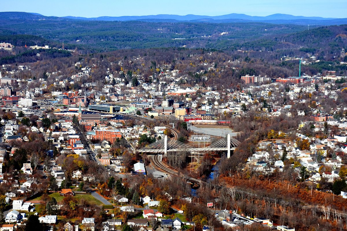 Aerial view of downtown Fitchburg in autumn, with the commuter rail tracks and a white cable bridge crossing the Nashua River valley below wooded hills