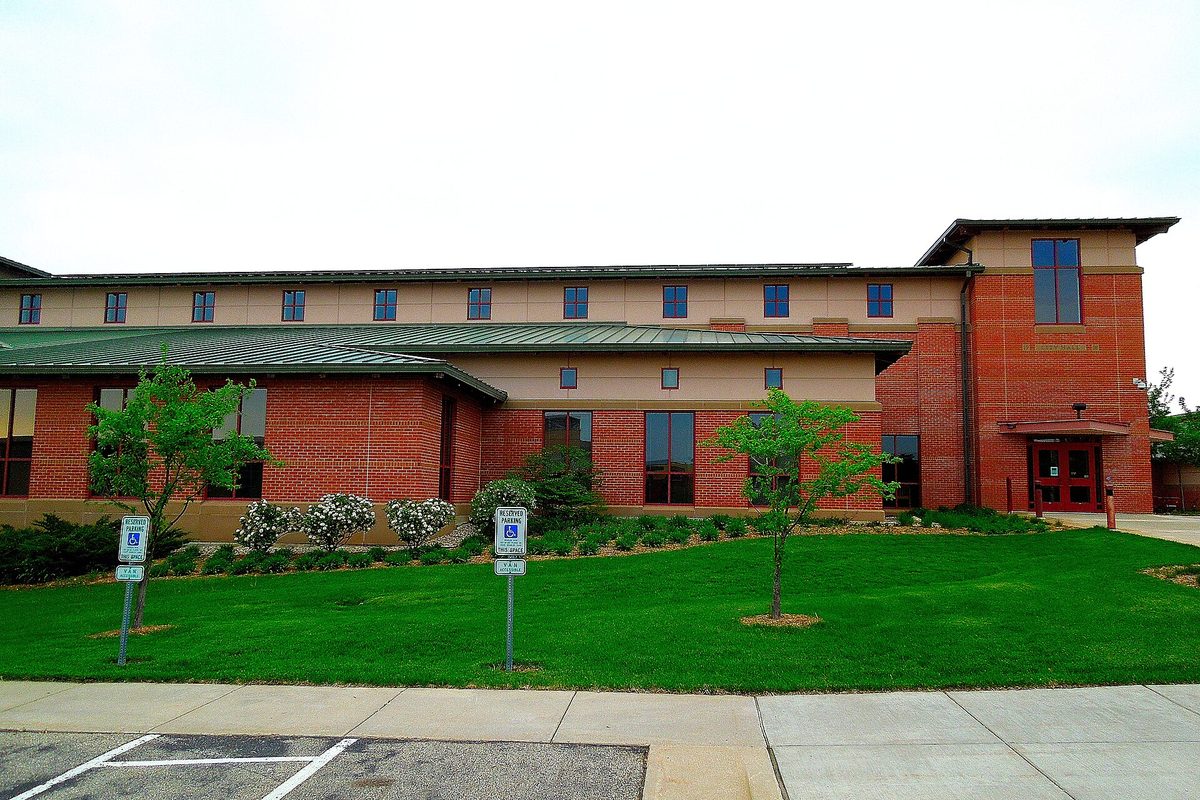 Fitchburg City Hall, a two-story brick and tan building with a green metal roof, set behind a mowed lawn, flowering shrubs and young trees