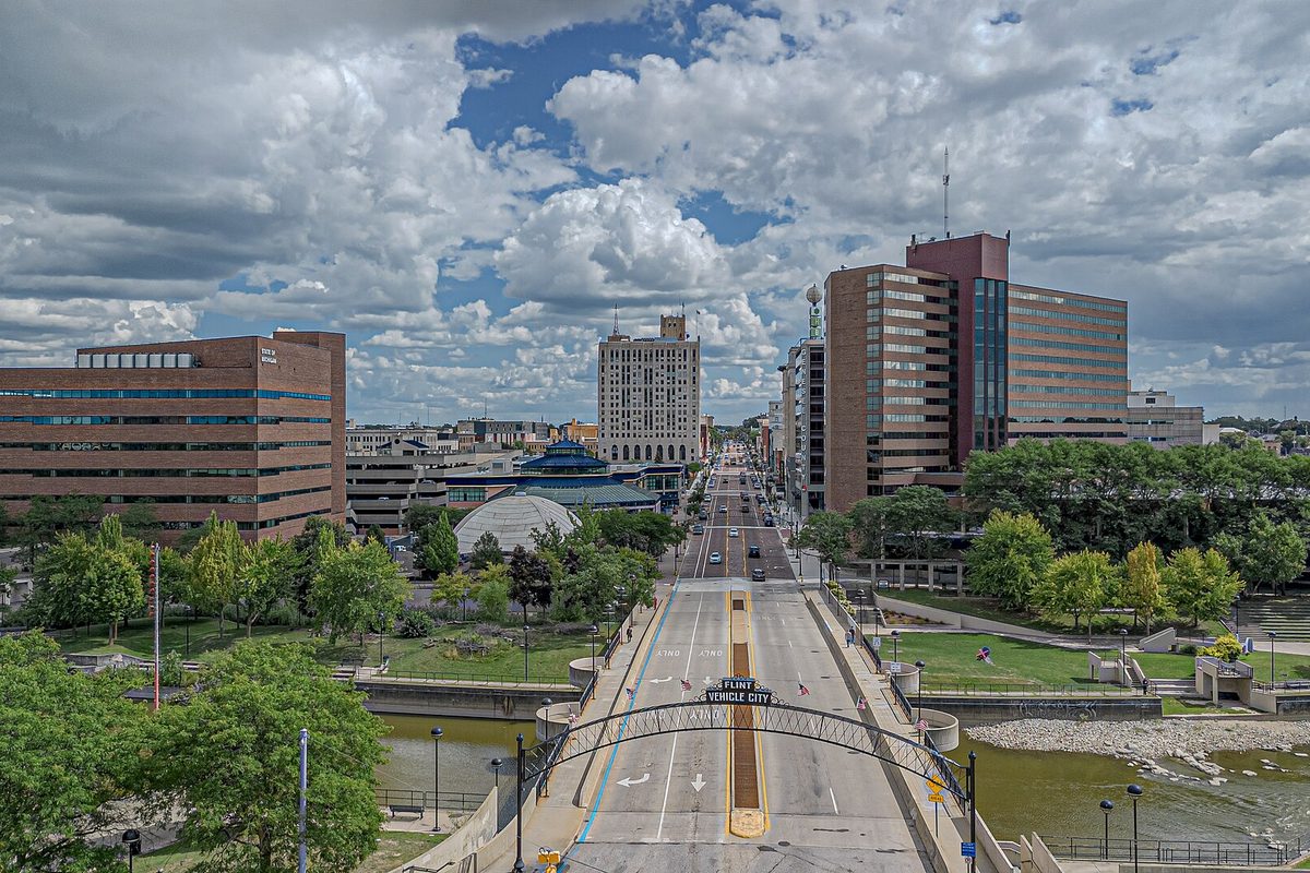 Aerial view of downtown Flint, with a main street running under a lettered arch and over a river, office towers and green parkland under a cloudy sky