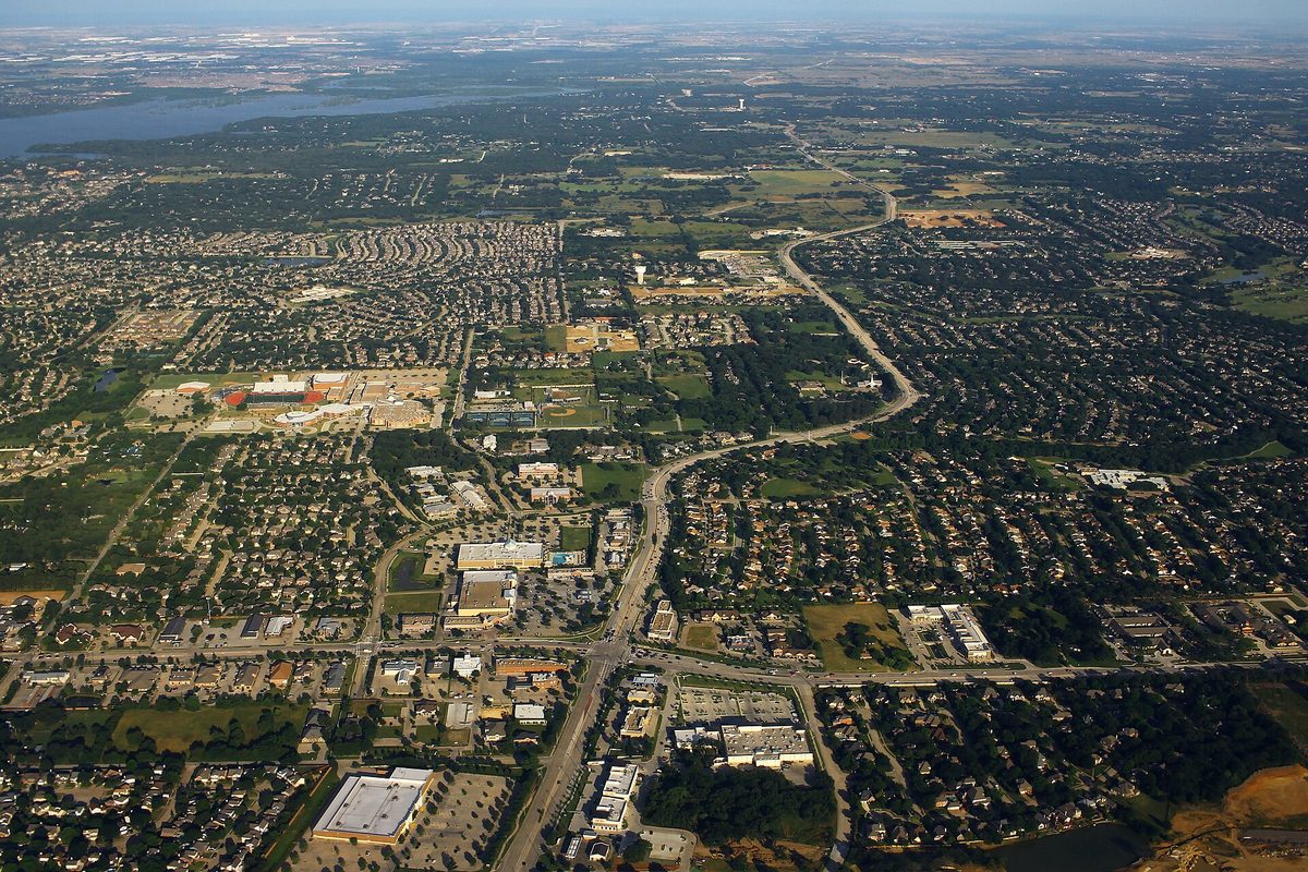Aerial view over Flower Mound: a divided road curving through neighborhoods and shopping centers, with a lake on the horizon