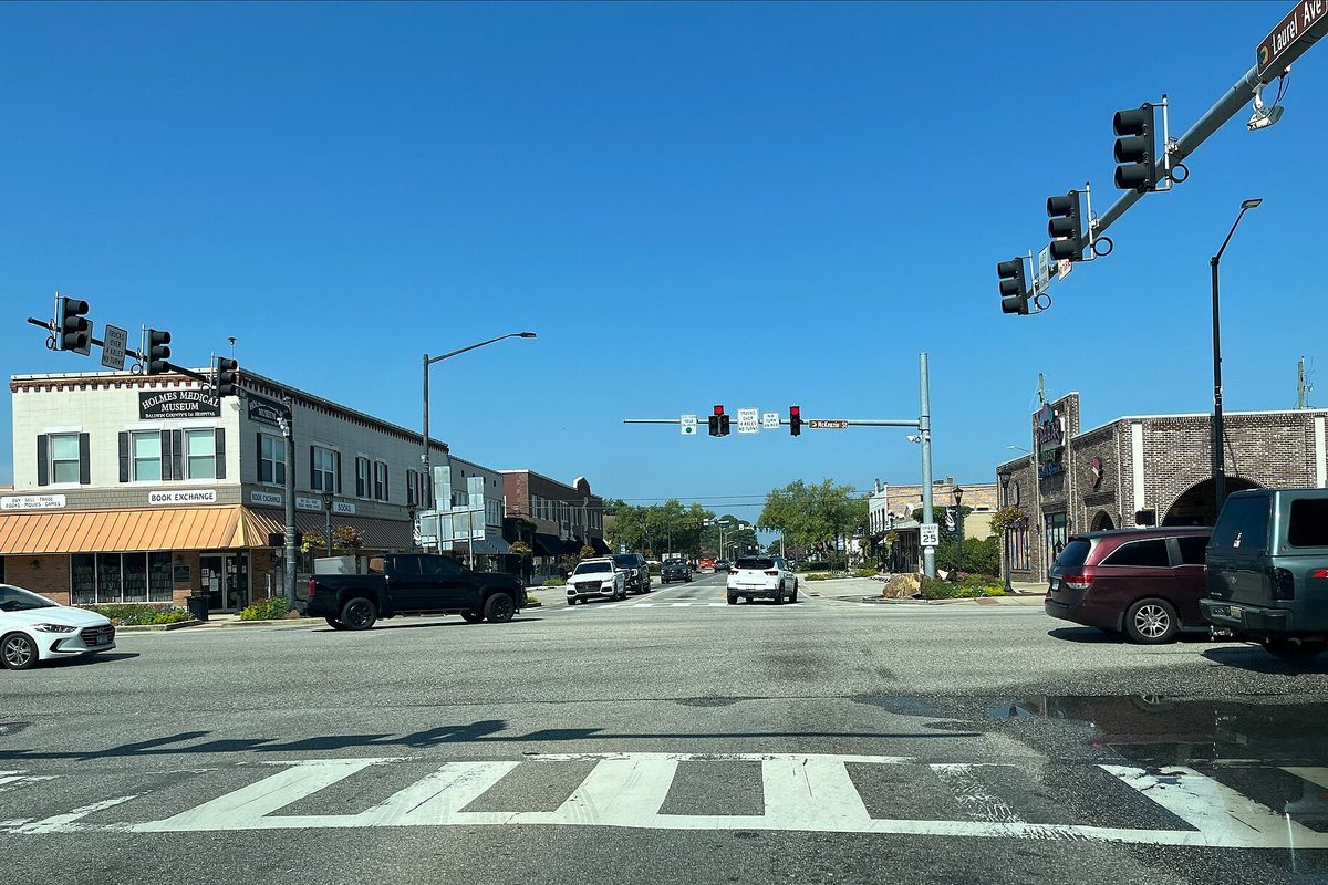 Traffic lights over the crossing of U.S. 98 and Alabama 59 in downtown Foley, with corner storefronts and a pickup waiting under a clear sky