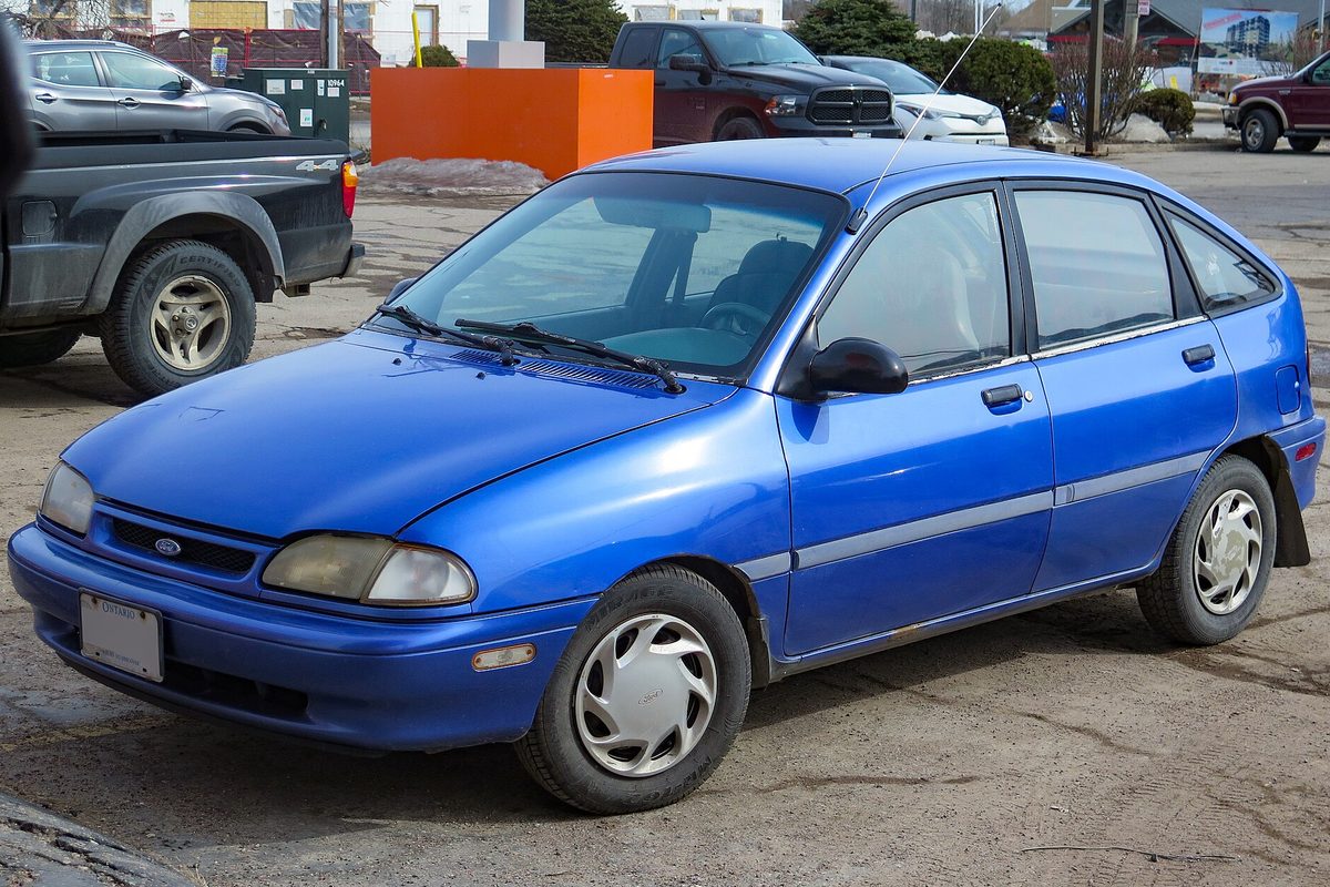 A bright blue 1994 Ford Aspire five-door hatchback with white wheel covers on a muddy gravel lot beside a gray pickup