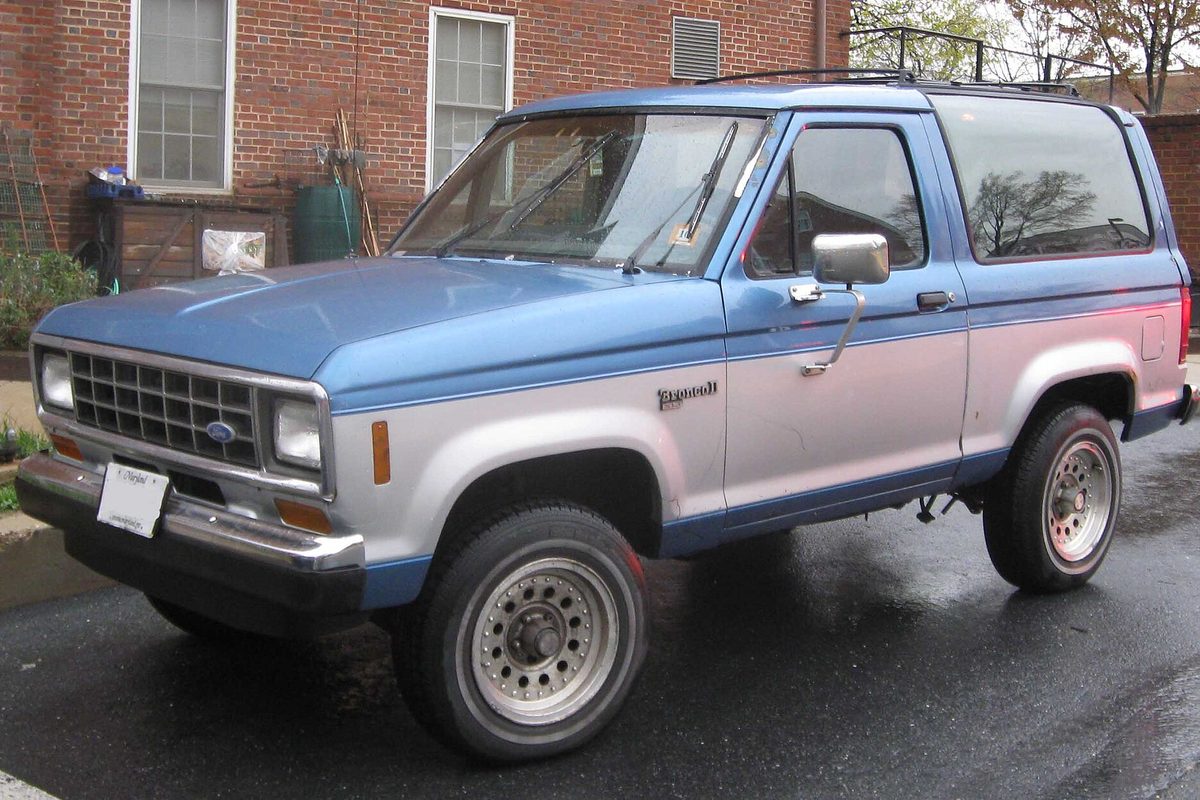 A blue-and-silver two-tone Ford Bronco II parked on a wet street in front of a brick apartment building