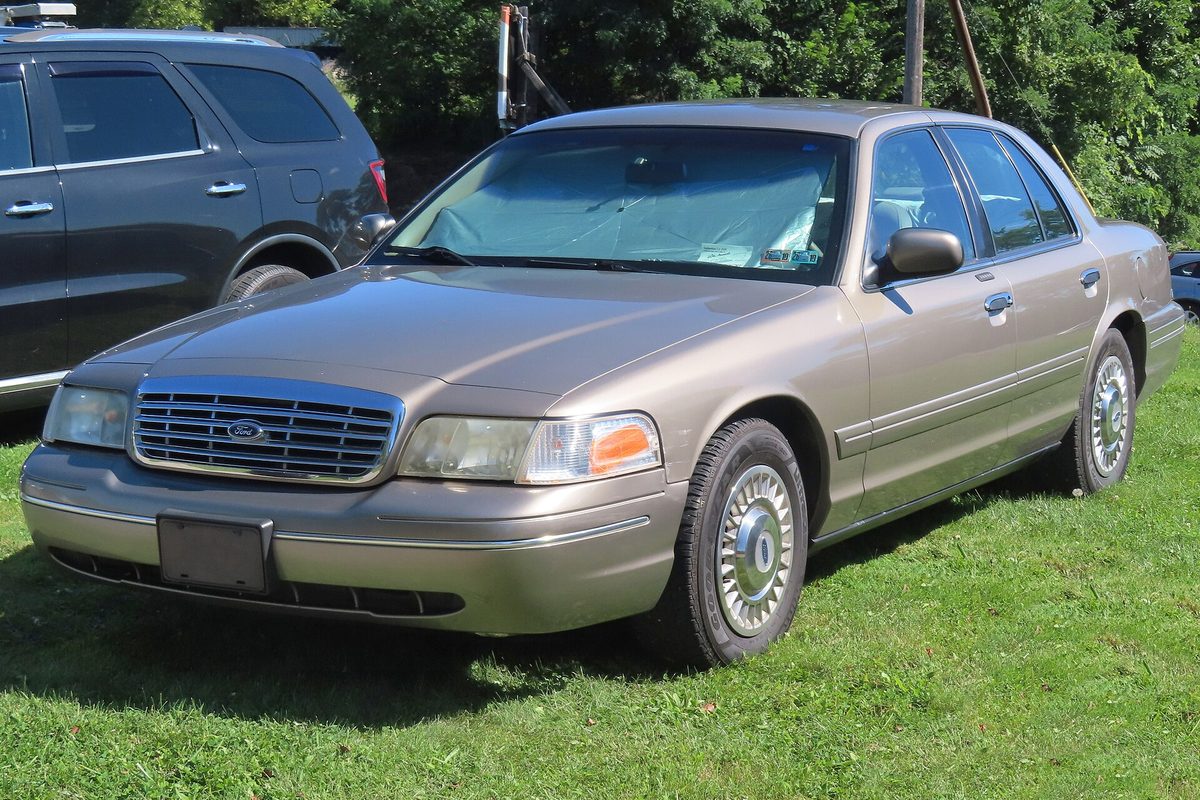 A beige 2001 Ford Crown Victoria with wire-style hubcaps and a sunshade in the windshield, parked on a grassy lot