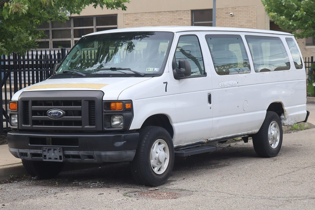A white 2011 Ford E-250 extended van with small dents along its side, parked at a curb beside a black iron fence