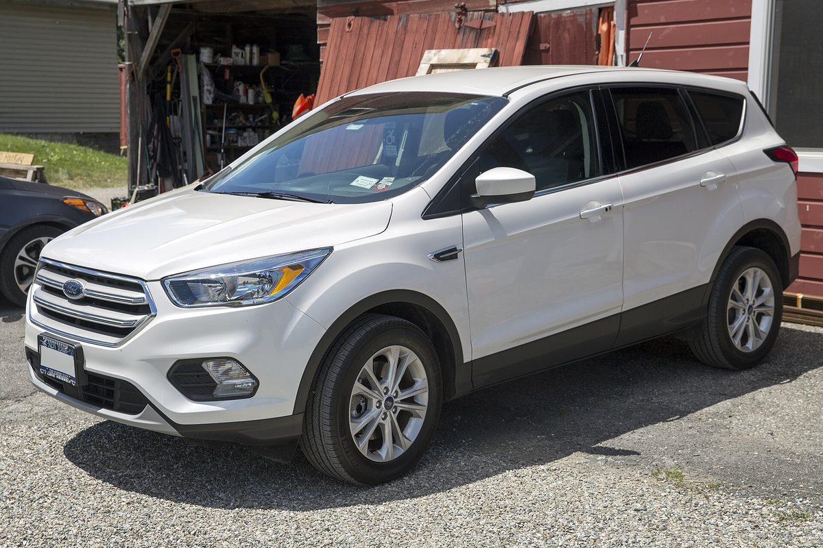 A white 2019 Ford Escape SE parked on gravel beside a red wooden building and an open tool shed