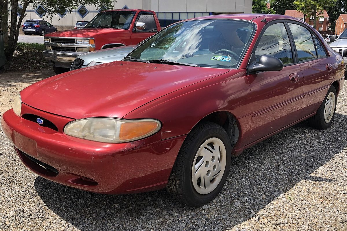 A red 1997 Ford Escort LX sedan with yellowed headlights parked on gravel, a red pickup truck behind it